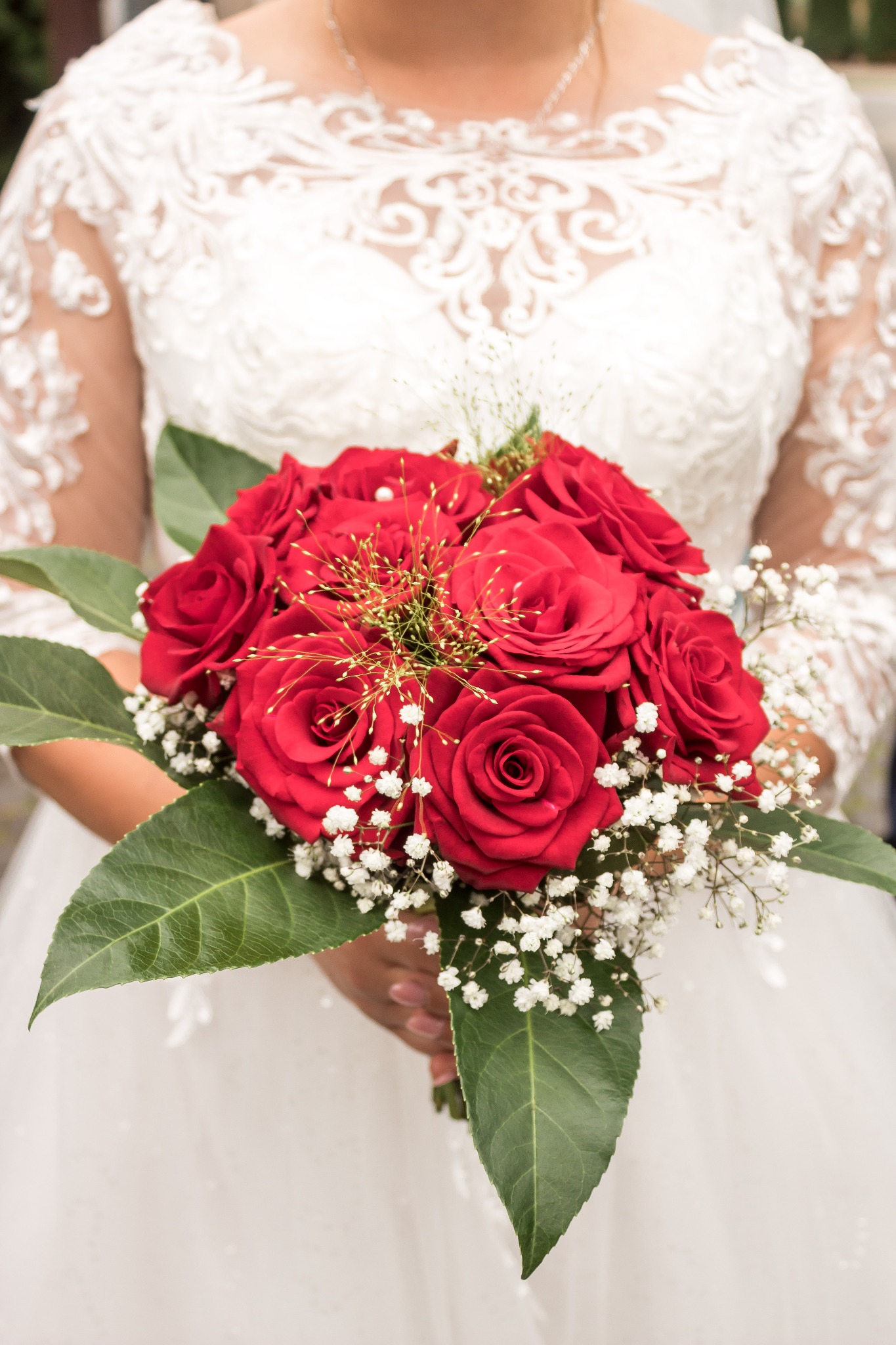Panna młoda w koronkowej sukni ślubnej trzyma bukiet z czerwonych róż i białych gipsówek, udekorowany złotymi elementami dekoracyjnymi.