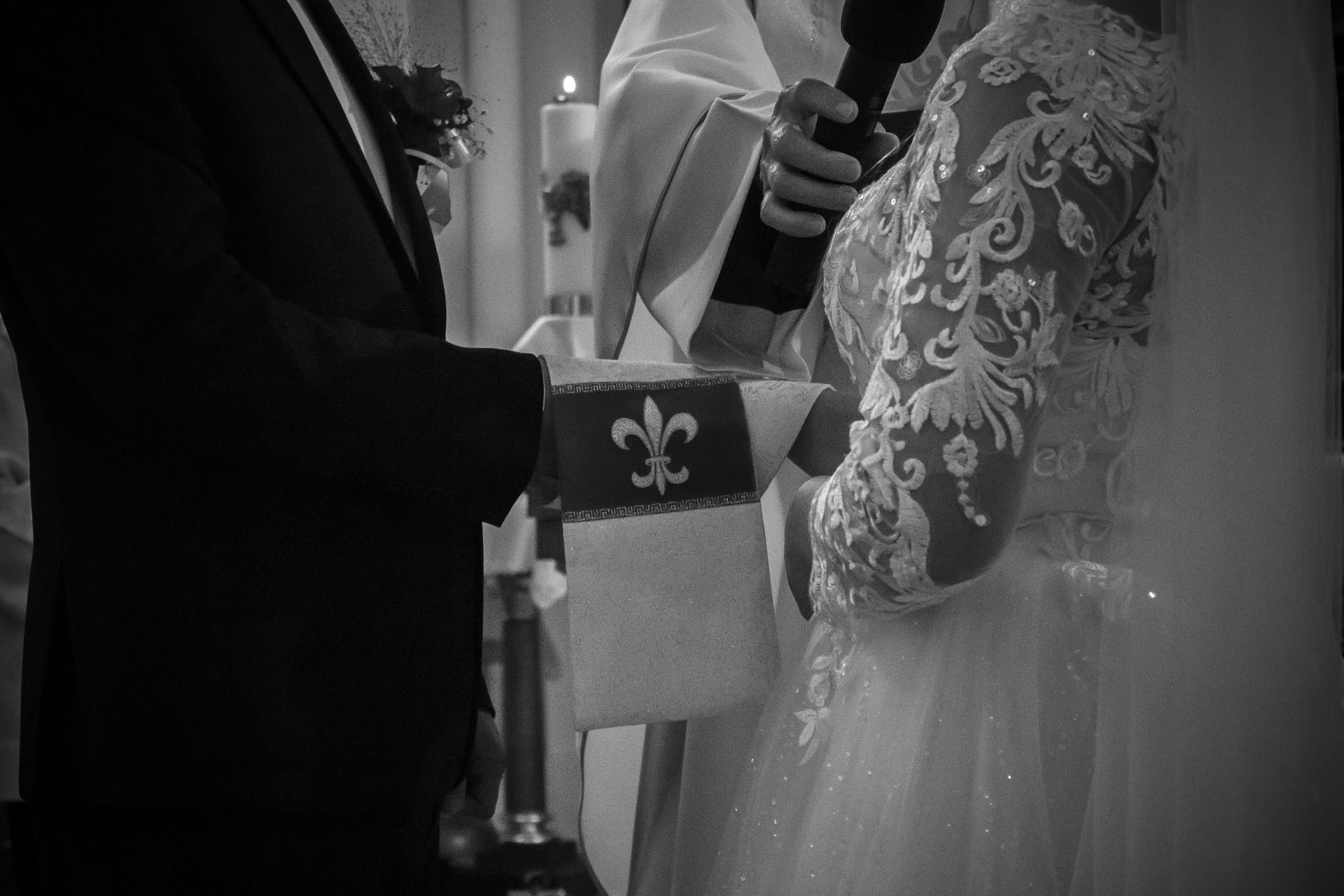 Czarno-białe zbliżenie dłoni nowożeńców podczas ceremonii ślubnej, ksiądz trzymający mikrofon w tle, widoczny fragment sukni ślubnej z koronkowymi zdobieniami i fragment garnituru pana młodego.