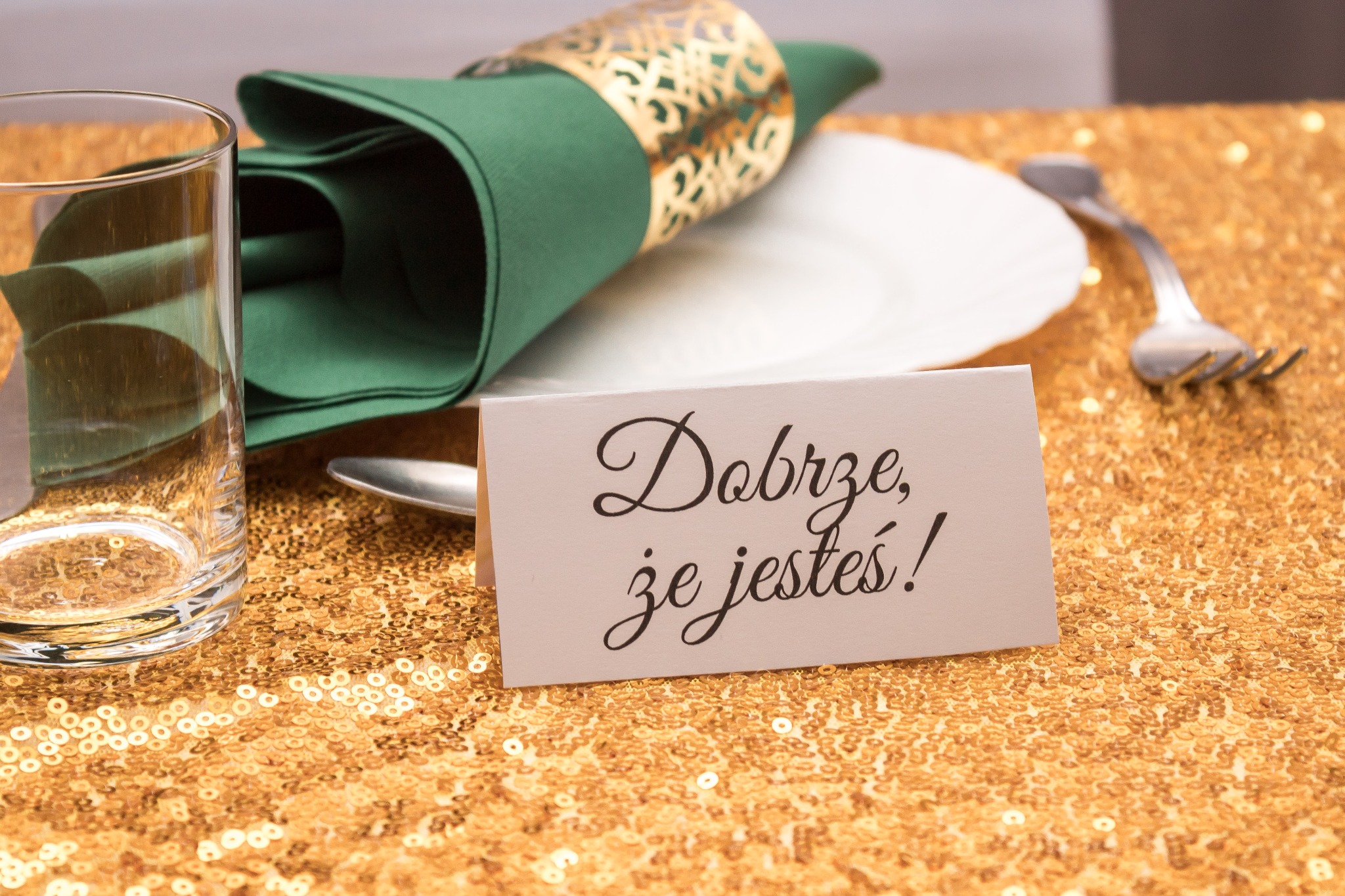 Elegancko nakryty stół z błyszczącym złotym obrusem, szklanką, zieloną serwetką w złotej obrączce, talerzem, sztućcami i kartką z napisem 'Dobrze, że jesteś!'