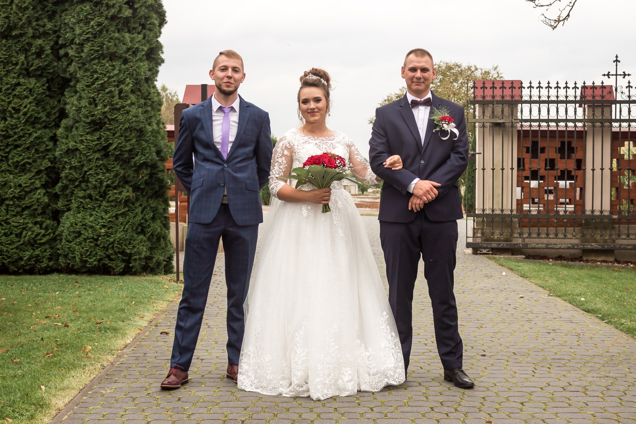 Panna młoda w białej sukni z bukietem czerwonych róż, otoczona dwoma mężczyznami w granatowych garniturach na brukowanej ścieżce przed ozdobną metalową bramą z krzyżem.