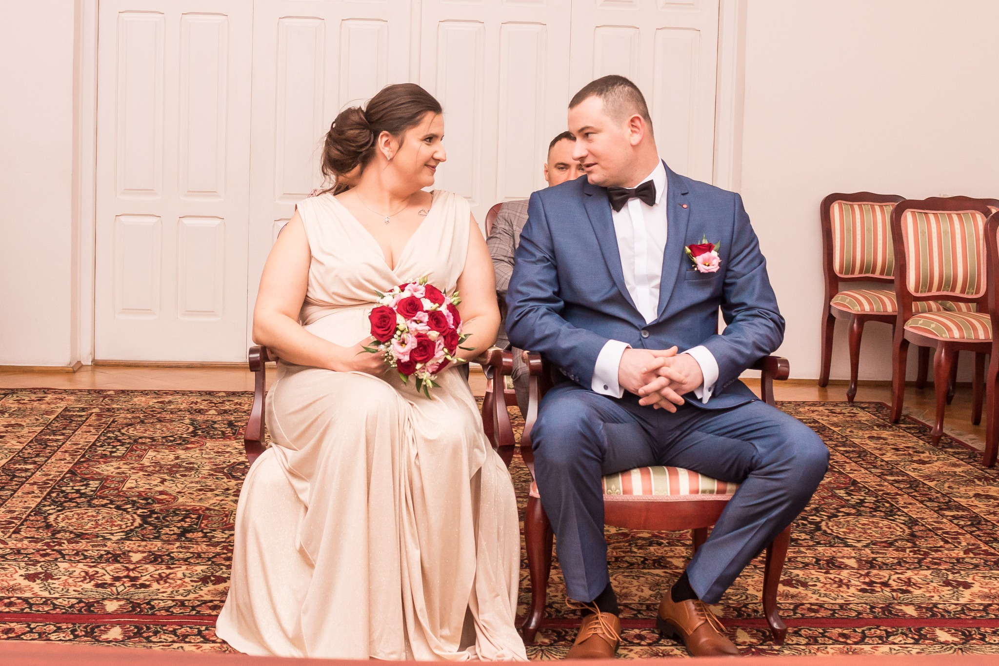 Para młoda siedzi na krzesłach podczas ceremonii, panna młoda trzyma bukiet czerwonych róż, pan młody w granatowym garniturze z muszką, w tle widoczne dodatkowe krzesła i fragment osoby.