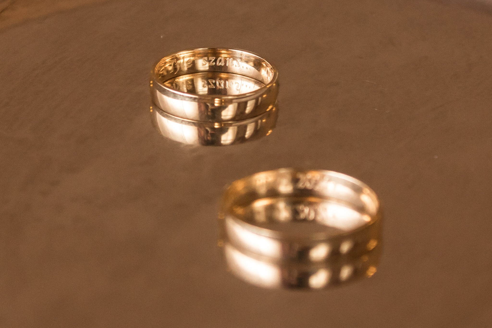 Dwa złote obrączki z wygrawerowanymi napisami, ustawione na błyszczącej, refleksyjnej powierzchni.