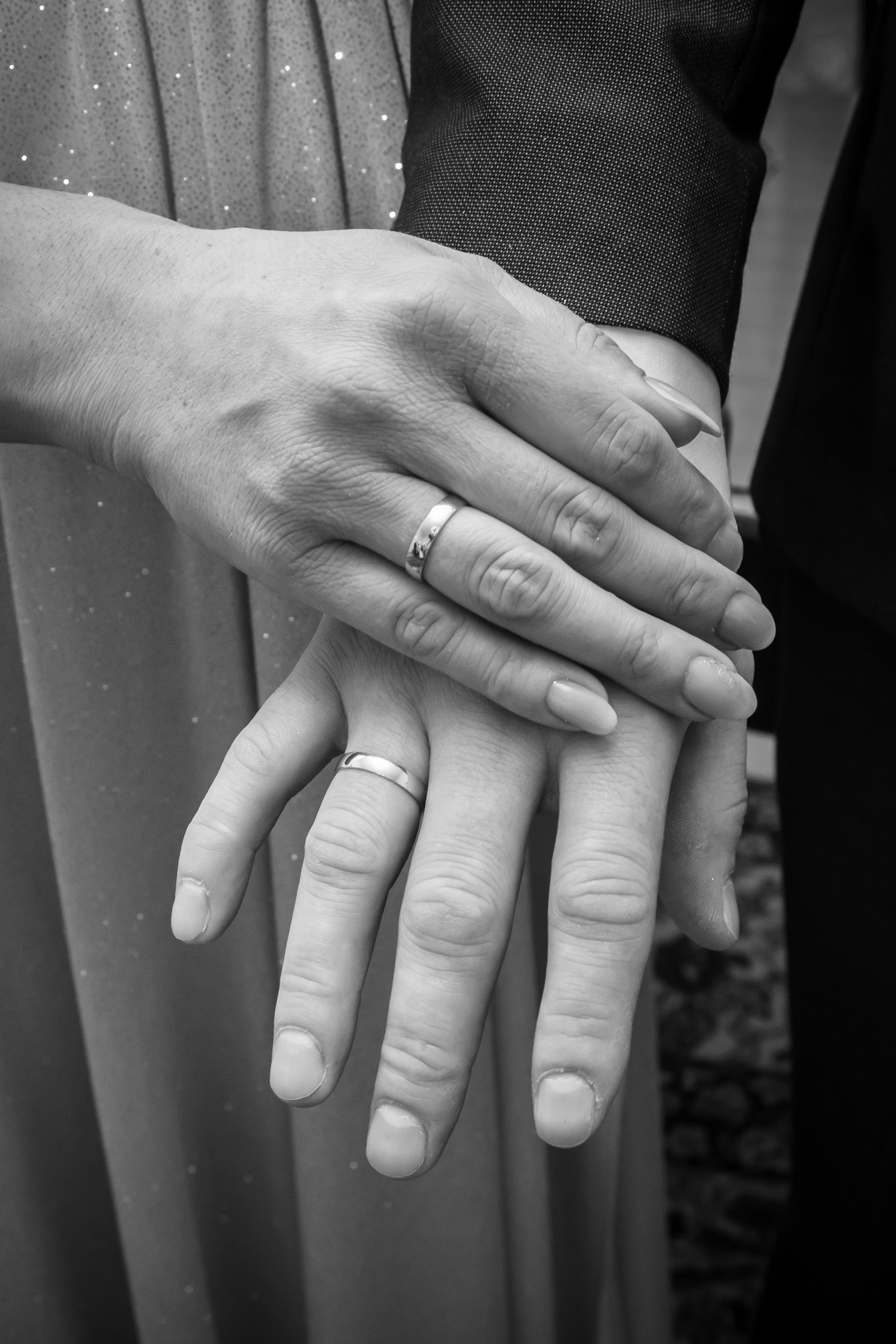 Dłonie pary z obrączkami ślubnymi, delikatnie splecione na tle sukienki z połyskującymi detalami i ciemnego garnituru.
