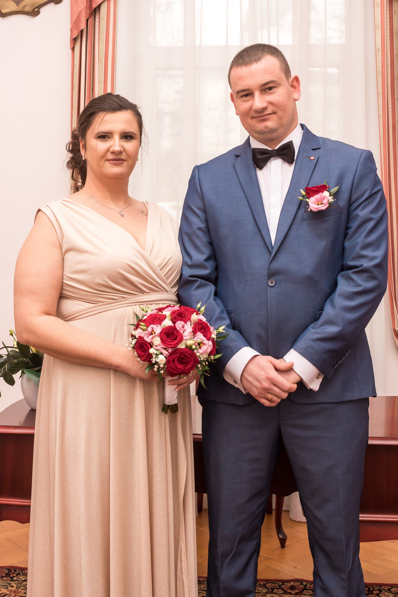 Para młoda: kobieta w długiej, beżowej sukni trzyma bukiet czerwonych i różowych róż, mężczyzna w niebieskim garniturze z muchą i butonierką z róż.