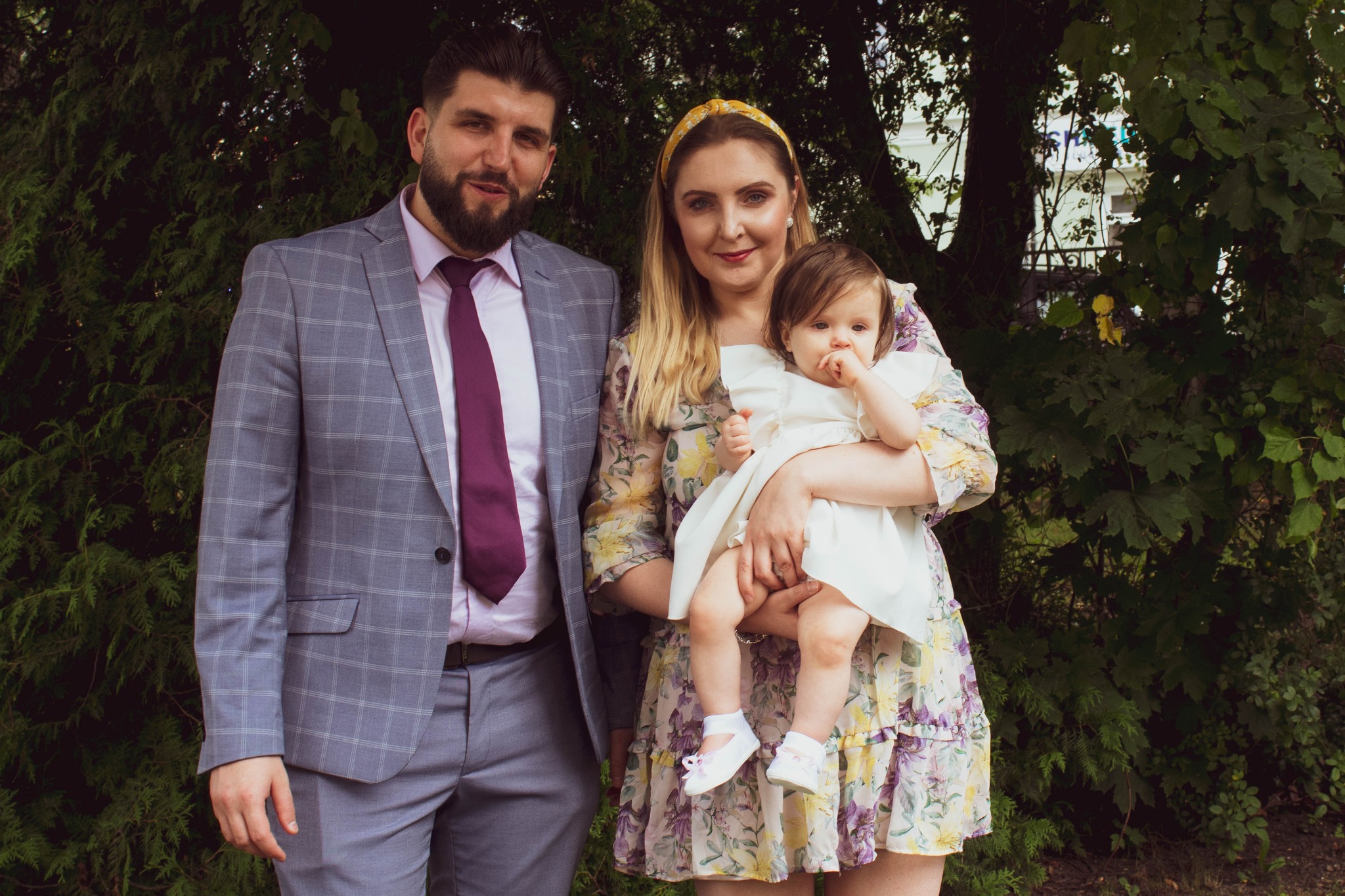 Rodzinne zdjęcie: Uśmiechnięty mężczyzna w szarym garniturze i fioletowym krawacie stoi obok kobiety w kwiecistej sukience trzymającej na rękach małą dziewczynkę w białej sukience i bucikach,...