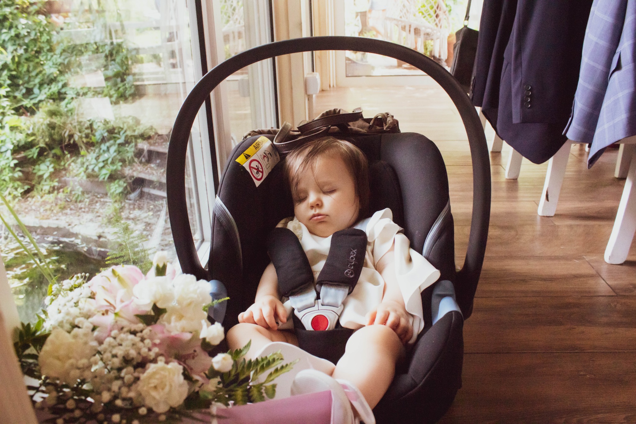 Śpiące niemowlę w foteliku samochodowym z bukietem kwiatów obok, w tle widok na zieleń z okna i wiszące marynarki.
