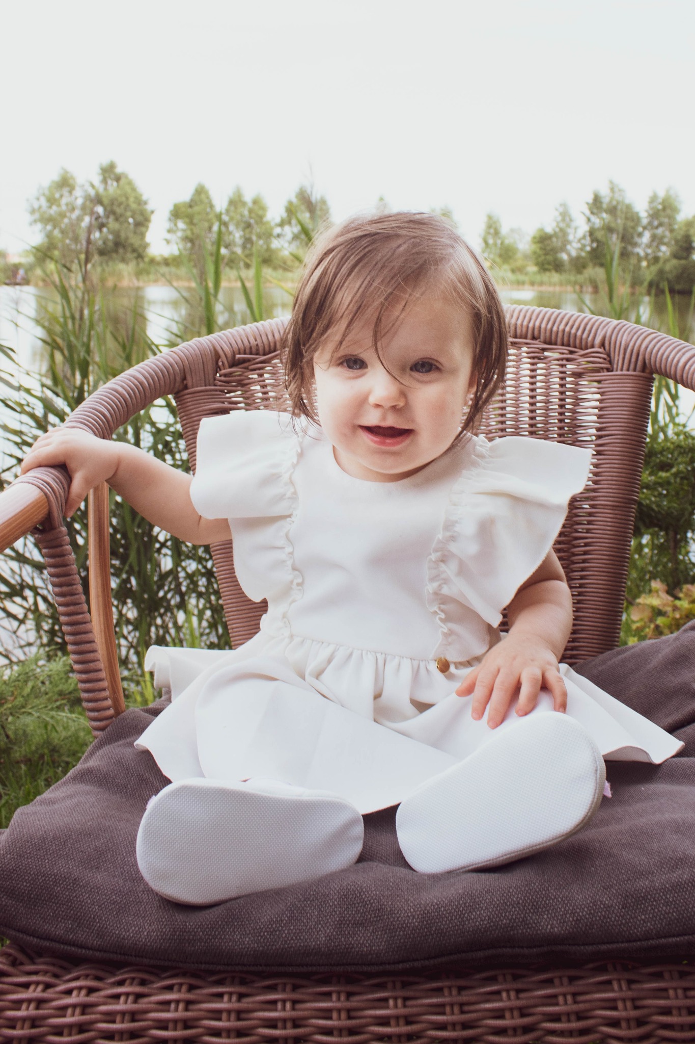 Uśmiechnięta dziewczynka w białej sukience siedzi w wiklinowym fotelu na tle jeziora i trzcin.