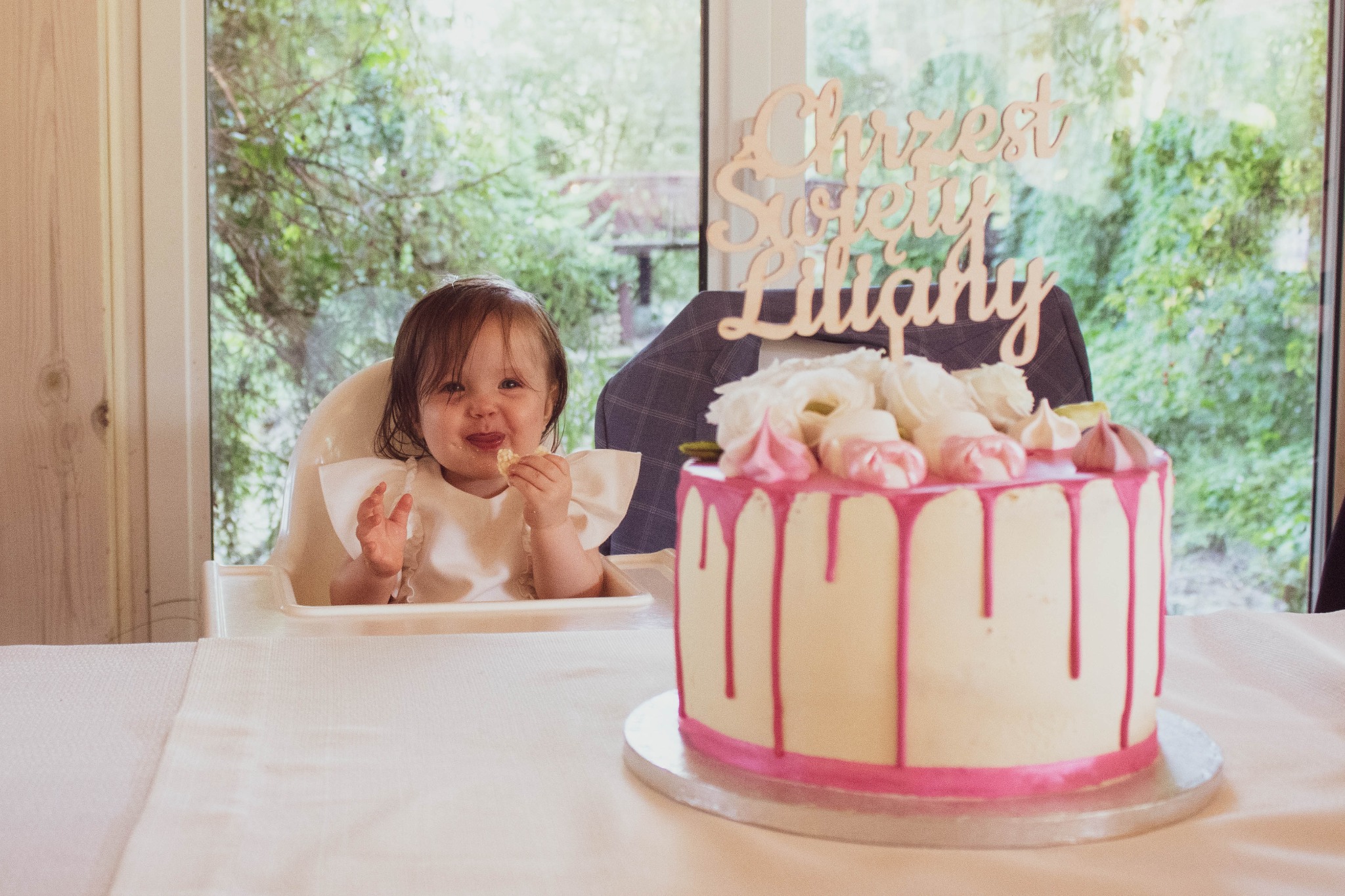 Roczne dziecko w krzesełku je ciastko, obok stoi tort z napisem 'Chrzest Święty Liliany' i dekoracją z białych kwiatów, za dzieckiem widok na zieleń przez okno.