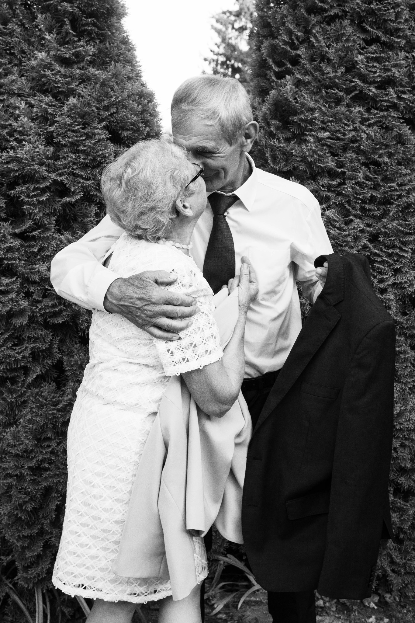 Czarno-białe zdjęcie starszej pary w objęciach na tle zielonych krzewów; mężczyzna w koszuli i krawacie całuje kobietę w sukience z koronki.