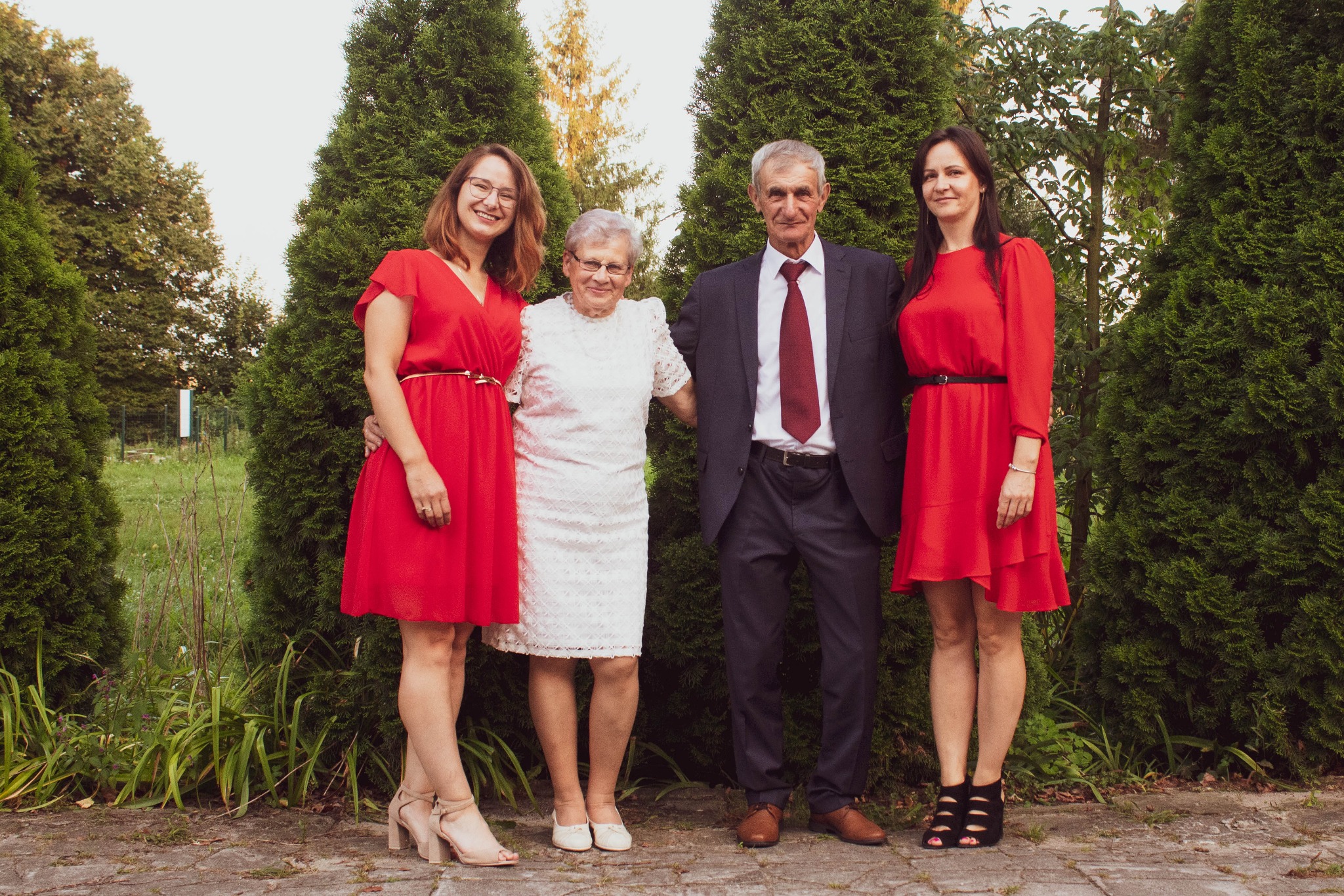 Rodzinne zdjęcie portretowe czterech osób: dwóch młodych kobiet w czerwonych sukienkach, starszej kobiety w białej sukience i mężczyzny w garniturze, stojących na tle zielonych krzewów.