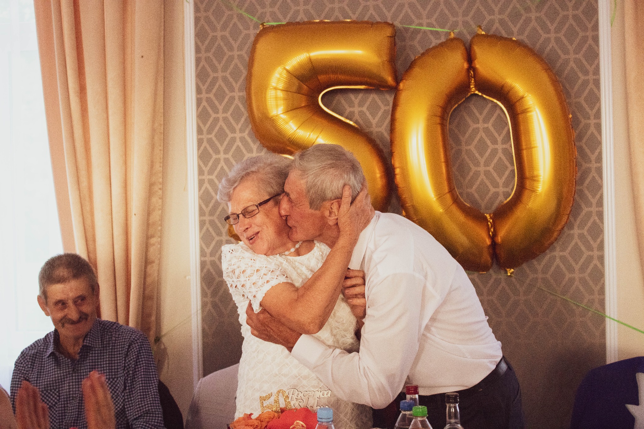 Para seniorów świętuje 50. rocznicę ślubu, obejmując się przed złotymi balonami w kształcie liczby 50 i tortem z napisem '50 rocznica'.
