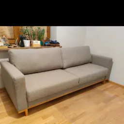 Szara, tapicerowana sofa z drewnianymi nóżkami na drewnianej podłodze, widok z przodu, za sofą okno z roślinami.