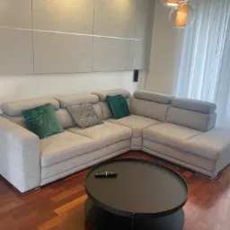 Szary narożnik z regulowanymi zagłówkami, zielonymi i szarą poduszką dekoracyjną, okrągły stolik kawowy na drewnianej podłodze, minimalistyczne panele ścienne i zwiewne zasłony w tle.