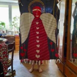 Osoba trzyma w górze duży, ręcznie szyty panel dekoracyjny przedstawiający stylizowanego anioła z czerwonymi sercami, widoczne stopy osoby i fragment wnętrza z drewnianą podłogą i szafą.