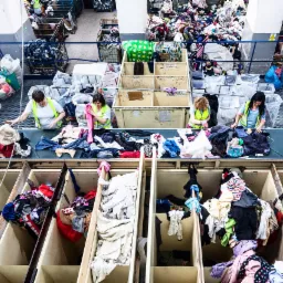Pracownicy sortują odzież na linii produkcyjnej w magazynie, widoczne pojemniki z ubraniami i górą składowane sterty ubrań.