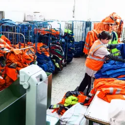 Kobieta w kamizelce odblaskowej sortuje niebieską odzież roboczą na stole w magazynie pełnym metalowych wózków z pomarańczową i niebieską odzieżą.