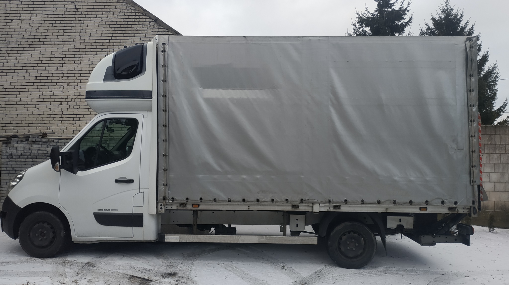 Biały samochód dostawczy z plandeką, widok z boku na tle muru z cegły i zaśnieżonego terenu.