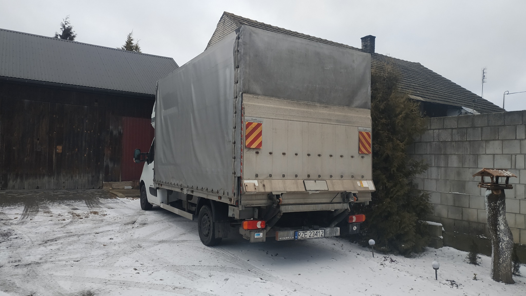Tył białego busa dostawczego z plandeką na podwórku pokrytym śniegiem, widoczne tablice ostrzegawcze i rejestracyjne
