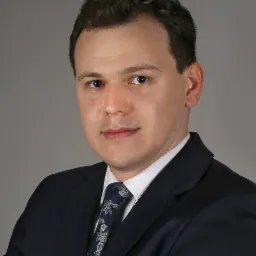 Portret mężczyzny w ciemnym garniturze i wzorzystym krawacie na szarym tle.