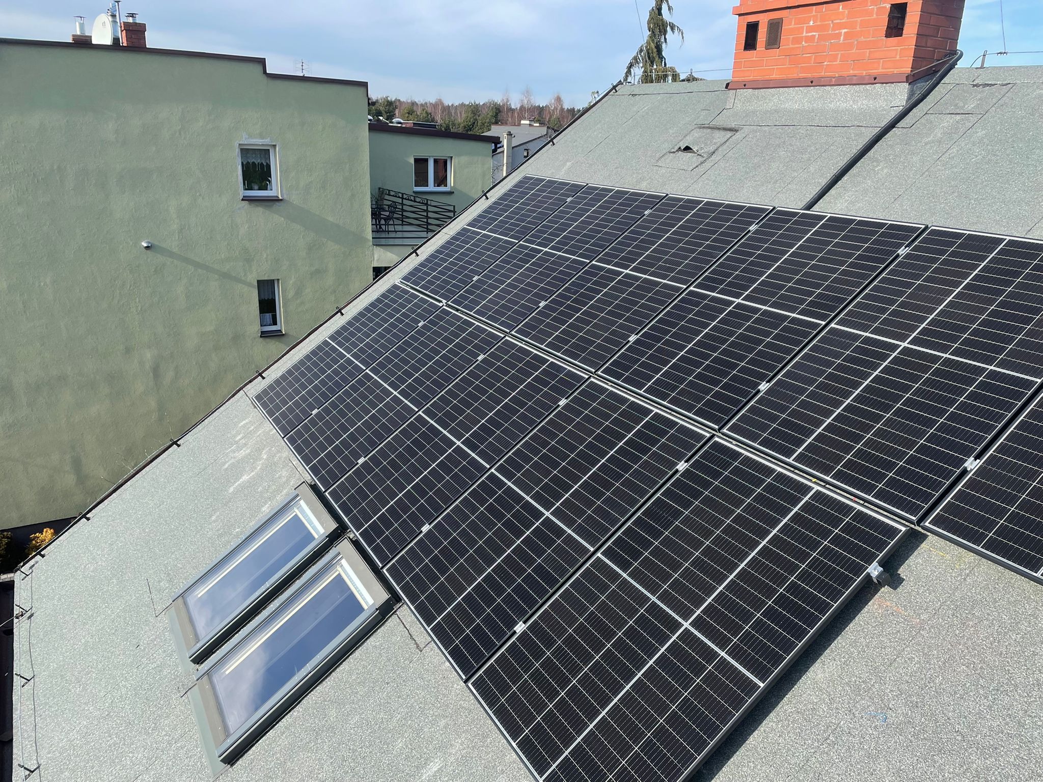Panele słoneczne zamontowane na dachu pokrytym papą, widoczne okna dachowe i komin z cegły, budynek w tle.