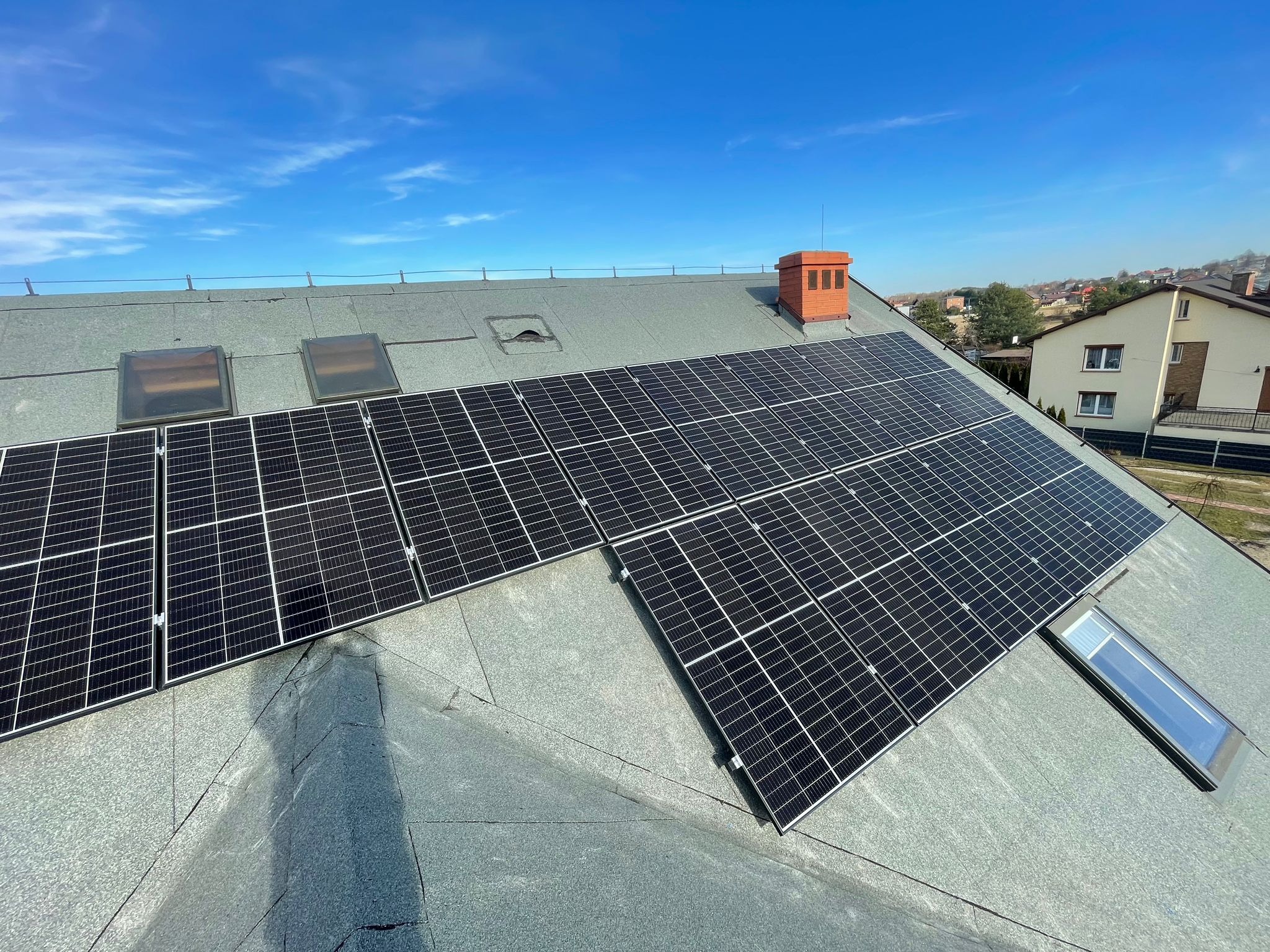 Panele słoneczne na płaskim dachu pokrytym papą, widoczne okna dachowe i komin wentylacyjny, niebieskie niebo w tle.