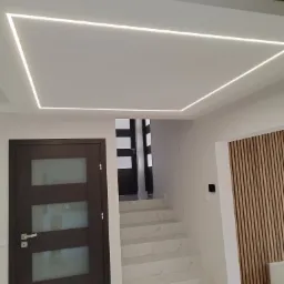 Nowoczesne wnętrze: białe schody z marmurowym wzorem, drewniana ściana z lamelami i podświetlany sufit z listwą LED tworzącą prostokąt.