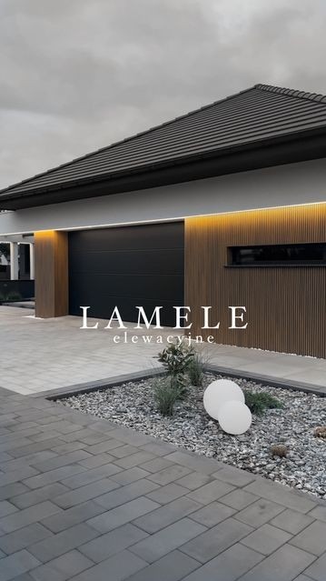 Nowoczesna elewacja domu jednorodzinnego z lamelami drewnianymi, oświetleniem LED i czarną bramą garażową, ozdobny ogródek z kamieniami i kulami świetlnymi.