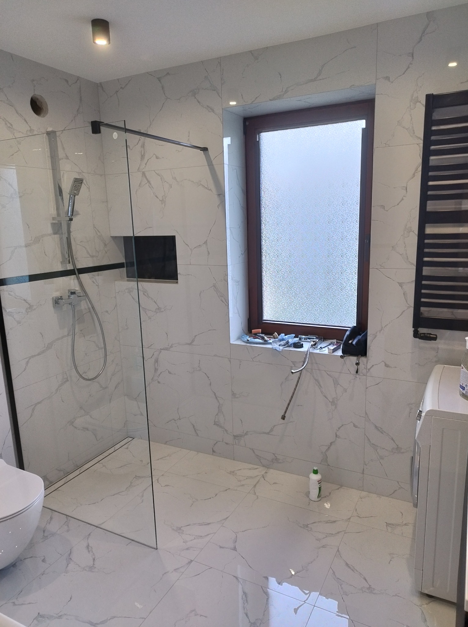 Nowoczesna łazienka w trakcie wykańczania: białe płytki imitujące marmur, szklana kabina prysznicowa typu walk-in, okno z narzędziami na parapecie i grzejnik drabinkowy.