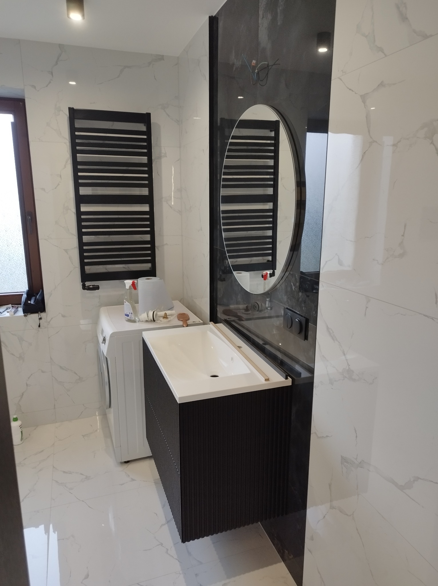 Wykończona łazienka z marmurowymi płytkami, czarnym grzejnikiem drabinkowym, owalnym lustrem, czarną szafką umywalkową z ryflowanym frontem i pralką z akcesoriami na górze.