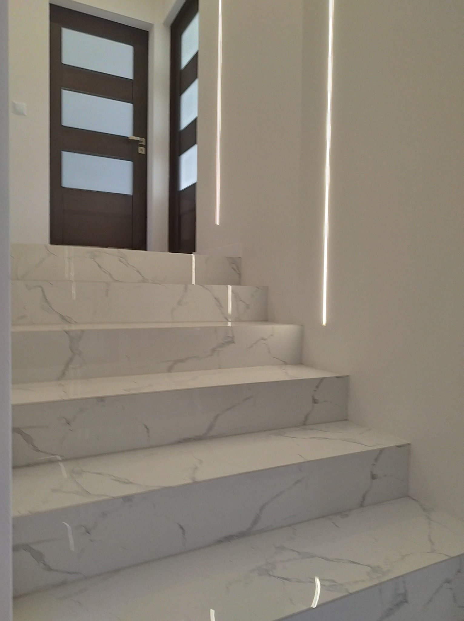 Schody wyłożone białym marmurem z szarymi żyłami, oświetlone pionowymi pasami światła LED na ścianie, z widocznymi drzwiami w tle.