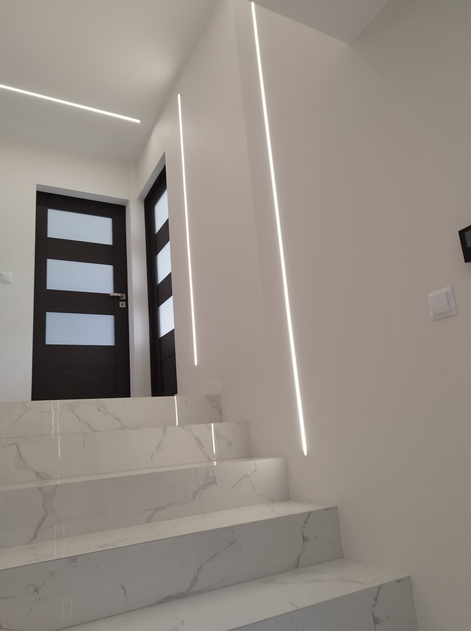 Schody wyłożone białymi płytkami imitującymi marmur, z podświetleniem LED na ścianach i suficie, oraz ciemne drzwi z szybami.