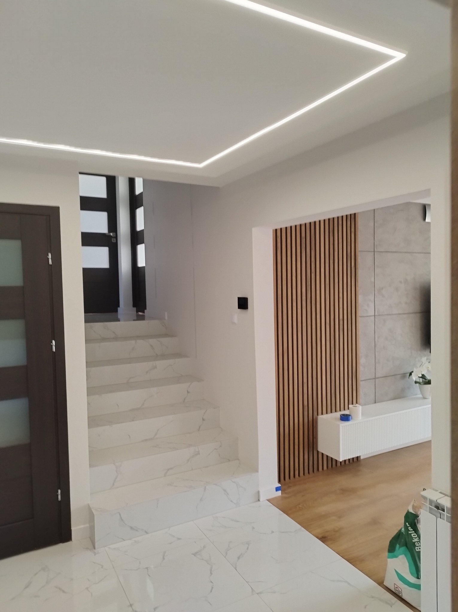 Nowoczesne wnętrze z marmurowymi schodami, podłogą i oświetleniem LED w suficie, widok na salon z drewnianą ścianką działową i białymi meblami.