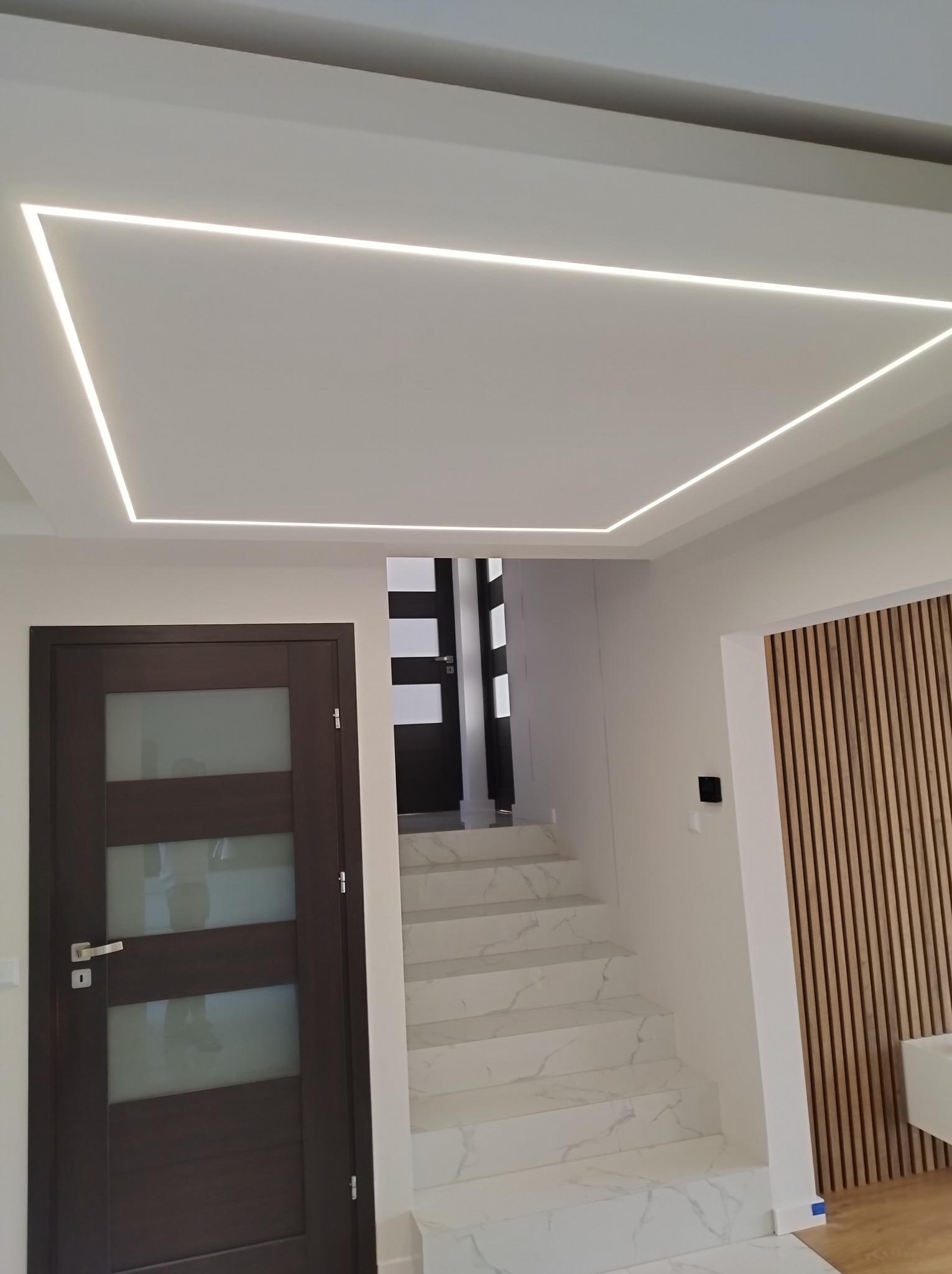 Nowoczesne wnętrze: białe schody z marmurowym wzorem, drewniana ściana z lamelami i podświetlany sufit z listwą LED tworzącą prostokąt.