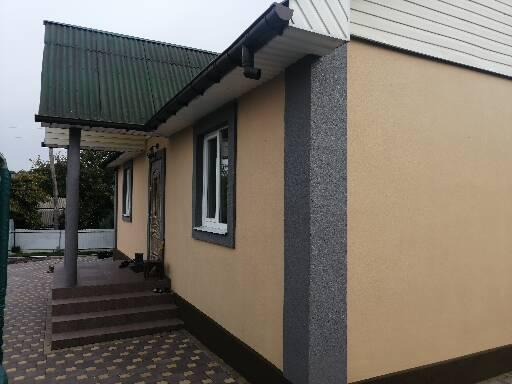 Wykończona elewacja domu jednorodzinnego z beżową strukturą tynku, ciemnoszarym narożnikiem, widoczne okno z białymi ramami, zielony dach i schody wejściowe z brązową kostką brukową.