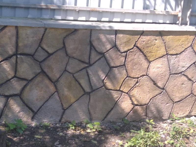 Fragment elewacji budynku z dekoracyjną okładziną imitującą kamienną ścianę z nieregularnych, brązowo-żółtych kamieni oddzielonych czarną fugą, widoczne drobne rośliny przy podstawie ściany...