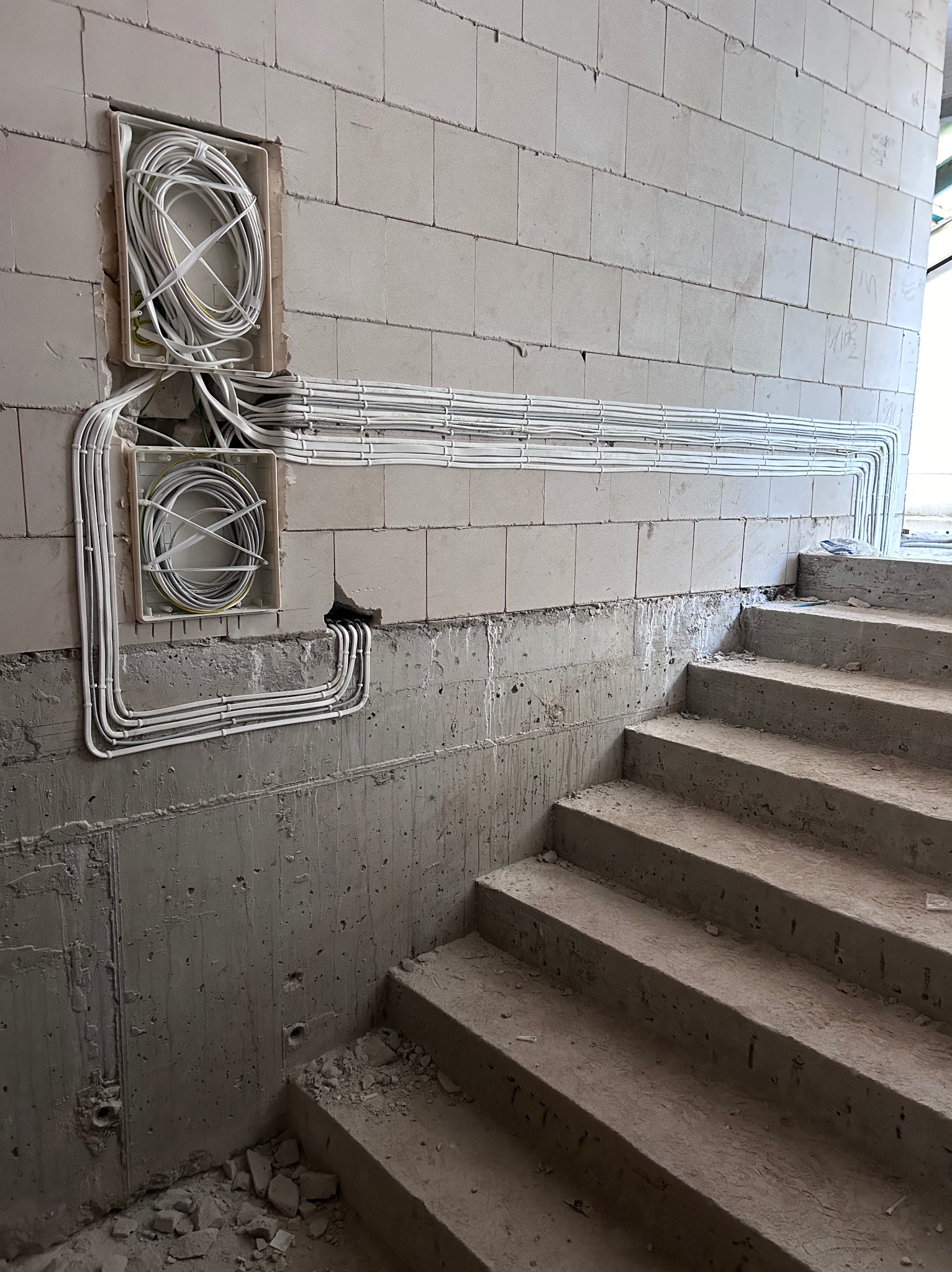 Białe przewody ułożone w równe rzędy na surowej ścianie z betonu komórkowego, prowadzące do dwóch puszek instalacyjnych. Widoczne schody w tle.