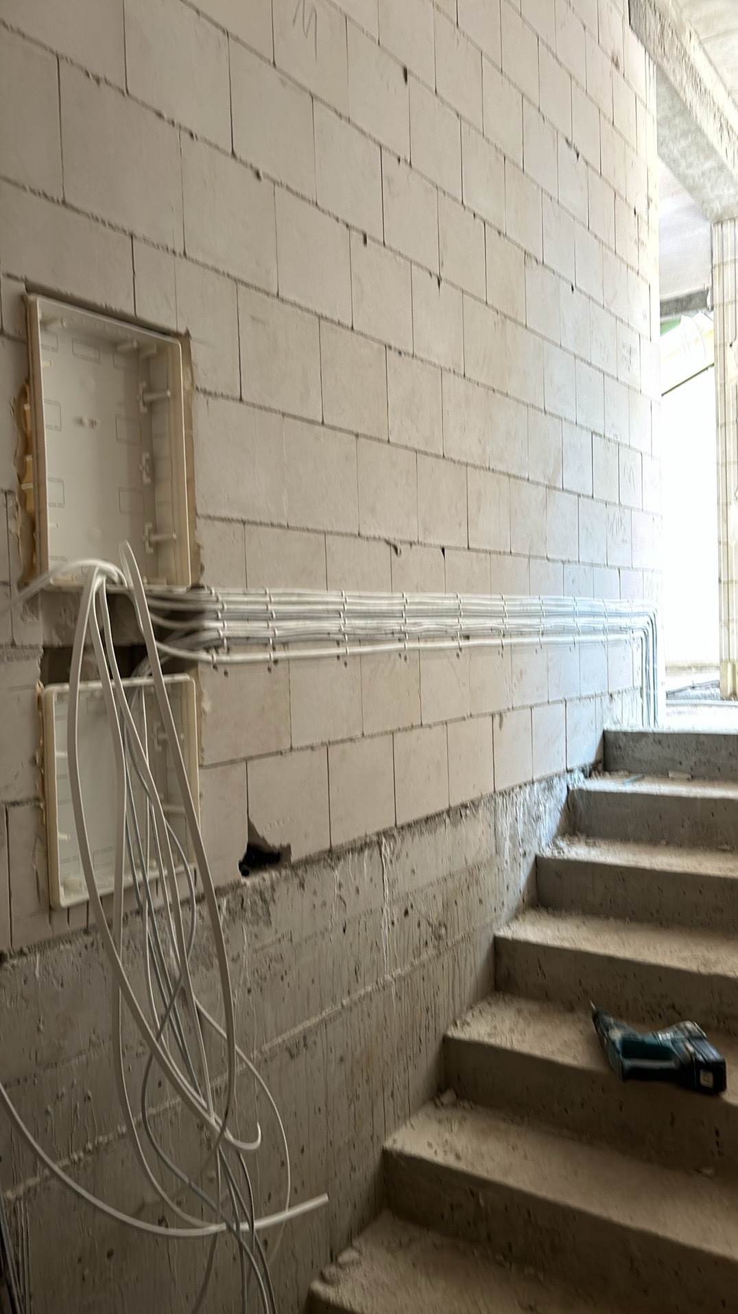 Instalacja przewodów w korytach kablowych, mocowanych do ściany z betonu komórkowego. Widoczne puszki instalacyjne i fragment schodów. Na schodach leży wiertarka.