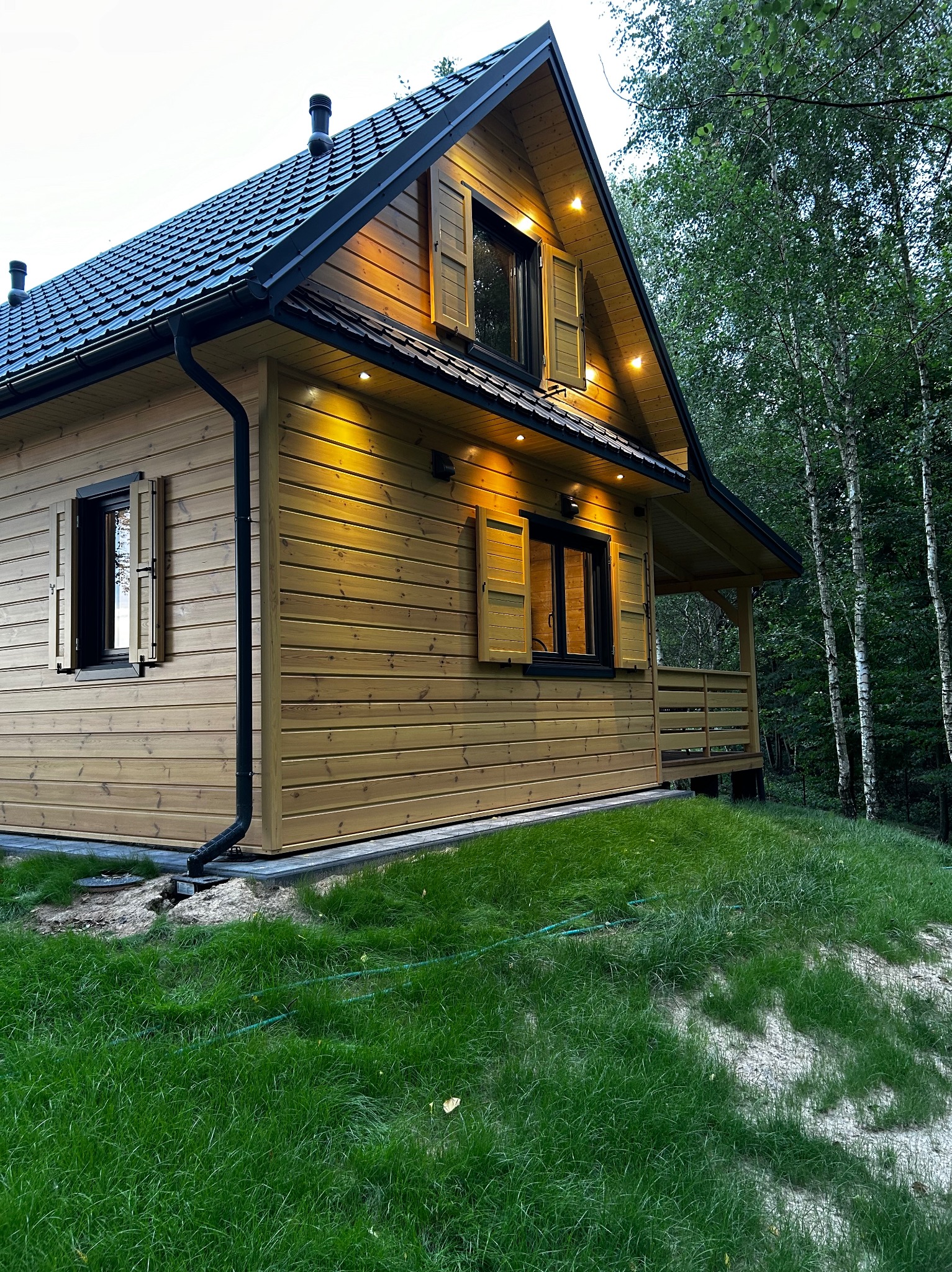 Elewacja drewnianego domu z czarną dachówką i oknami z okiennicami, oświetlona ciepłym światłem, na tle drzew. Widoczne rynny i fragment trawnika.