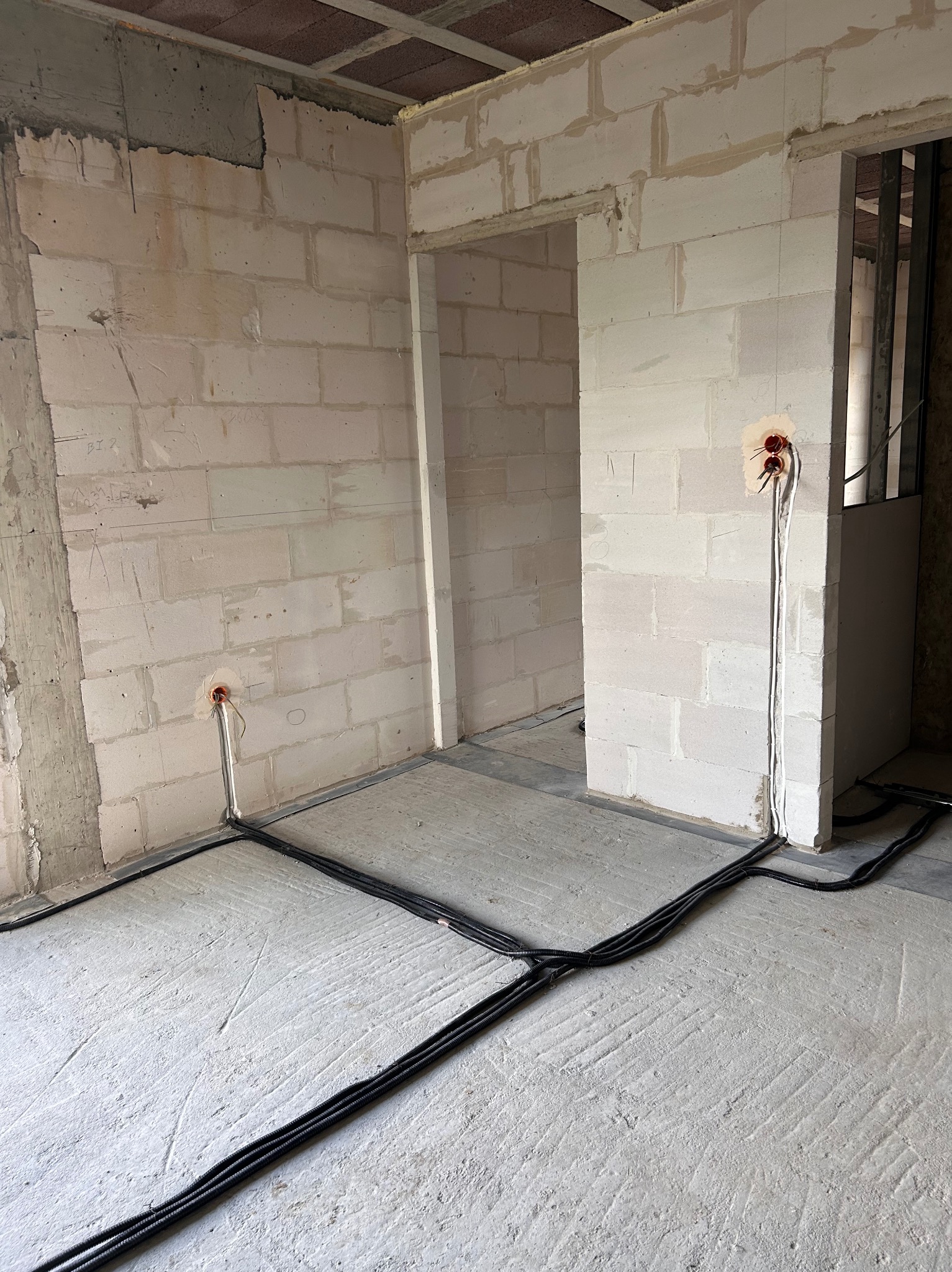 Surowy stan wnętrza z instalacją elektryczną: kable w czarnych peszlach ułożone na podłodze, wystające ze ściany przewody w puszkach. Ściany z białego betonu komórkowego.