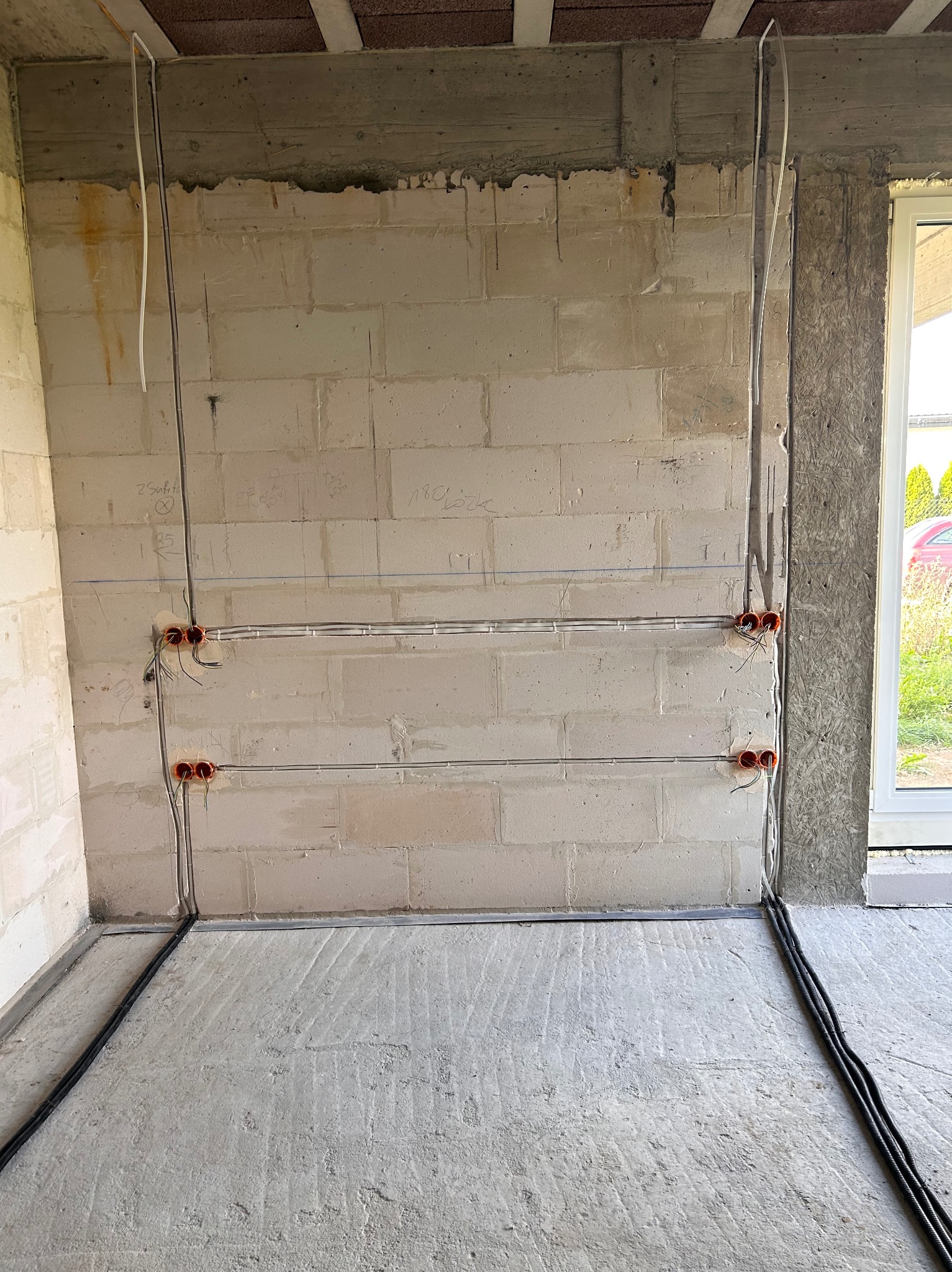 Surowy stan: instalacja elektryczna w nowym budynku, kable w peszlach przy podłodze, puszki podtynkowe w ścianie z betonu komórkowego, widok na okno i fragment zieleni.