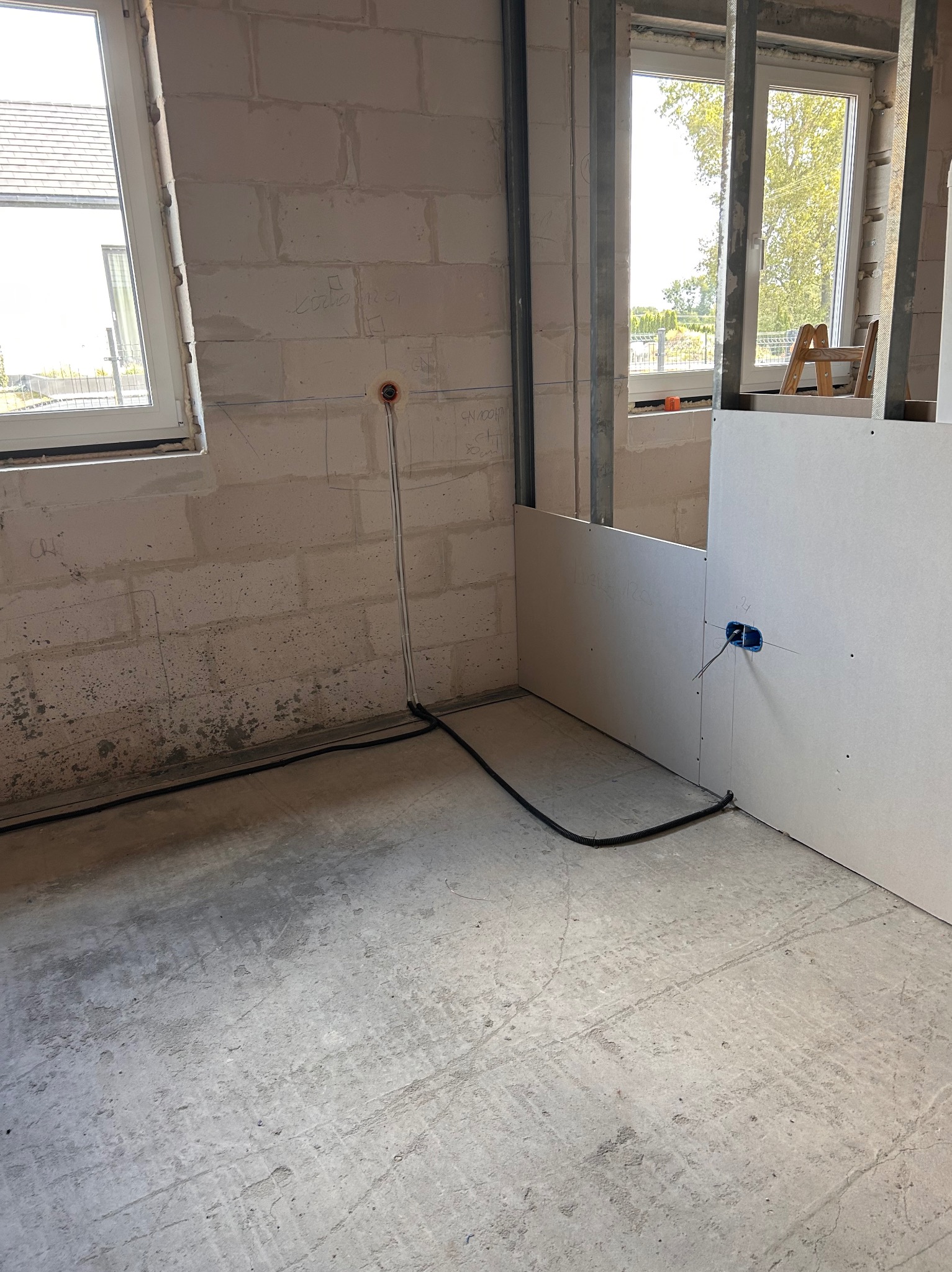 Wnętrze w trakcie wykończenia: ściany z pustaków, instalacja elektryczna w peszlu, płyty karton-gips na stelażu, okno z widokiem na zieleń. Prace remontowe w toku.