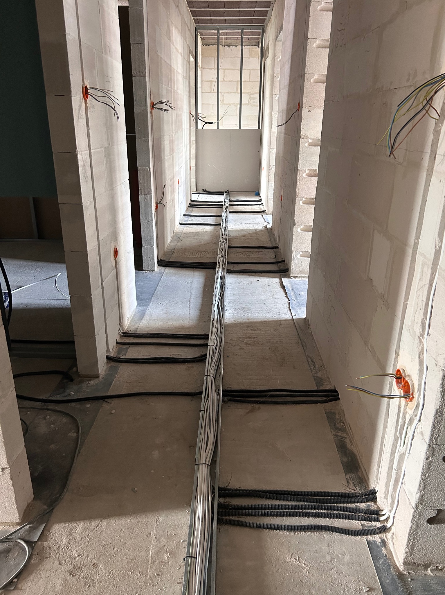 Wnętrze w trakcie prac elektrycznych: korytarz z instalacją kabli w peszlach i puszkami elektrycznymi w ścianach z bloków, surowy stan deweloperski.