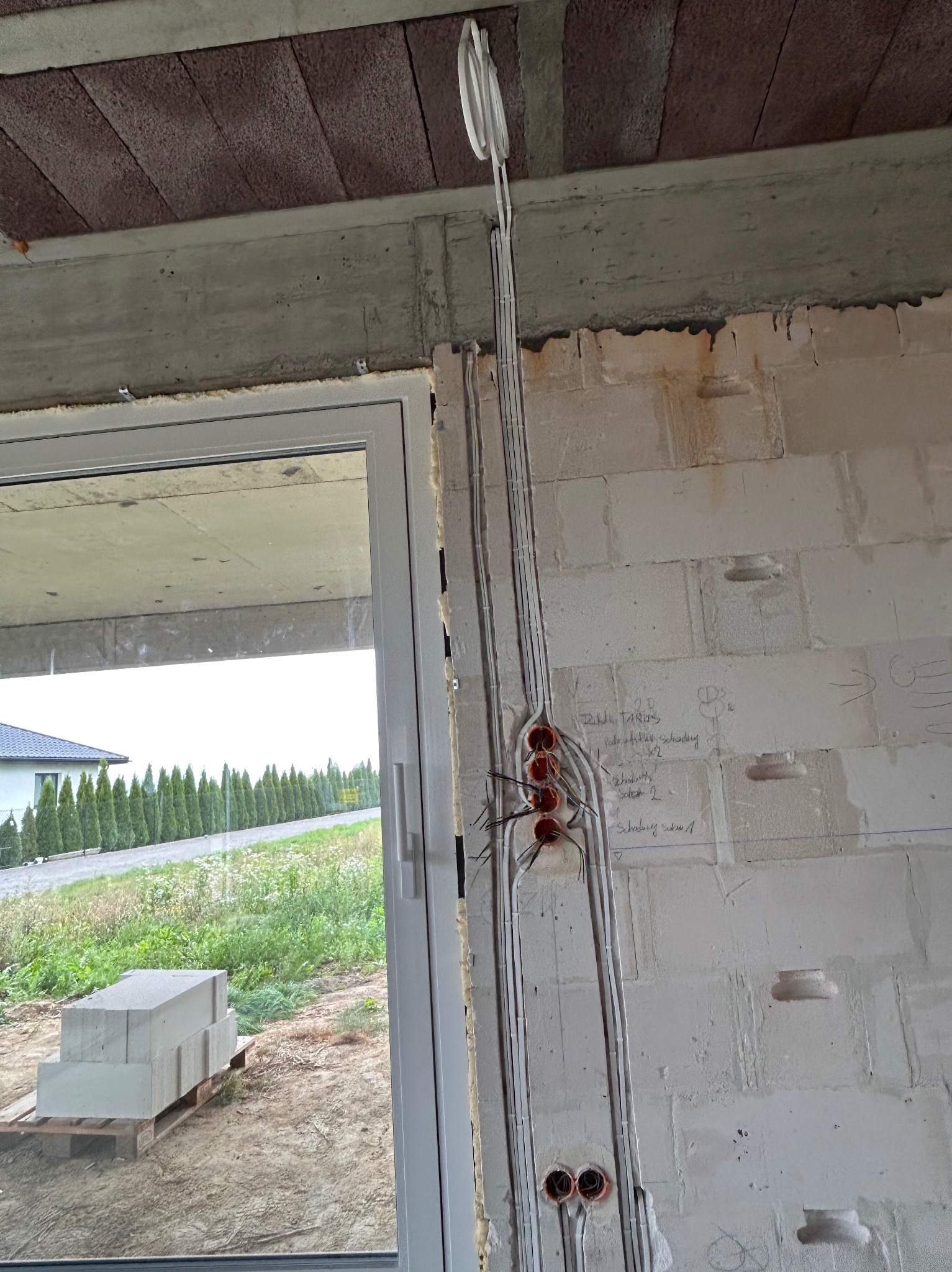 Surowe wnętrze w trakcie prac: widoczne kable elektryczne prowadzone po ścianie z betonu komórkowego, zarys instalacji przed tynkowaniem. Okno z widokiem na zieleń.