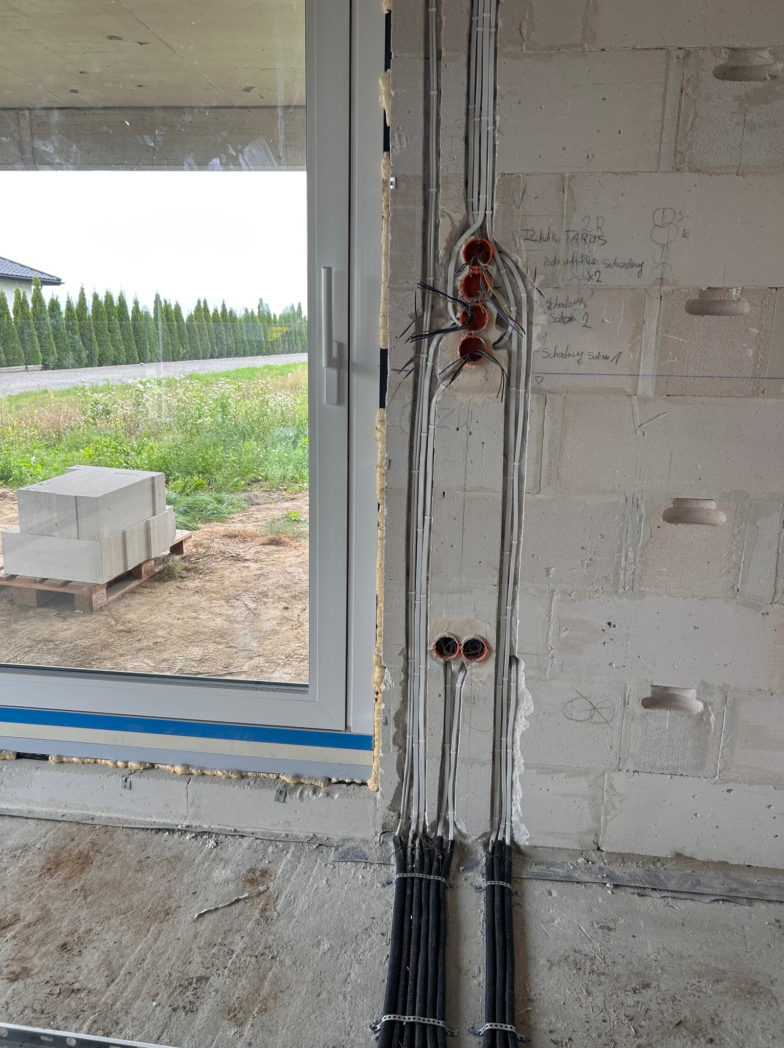 Surowa ściana z widocznymi przewodami w peszlach i puszkami instalacyjnymi. Widoczne napisy na ścianie i okno z widokiem na zewnątrz.