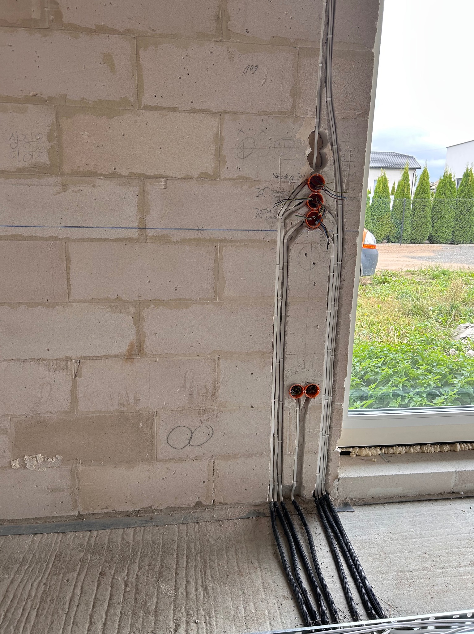 Ściana z instalacją elektryczną w trakcie montażu, widoczne kable w peszlach prowadzone pionowo. Na ścianie widoczne oznaczenia i puszki instalacyjne.