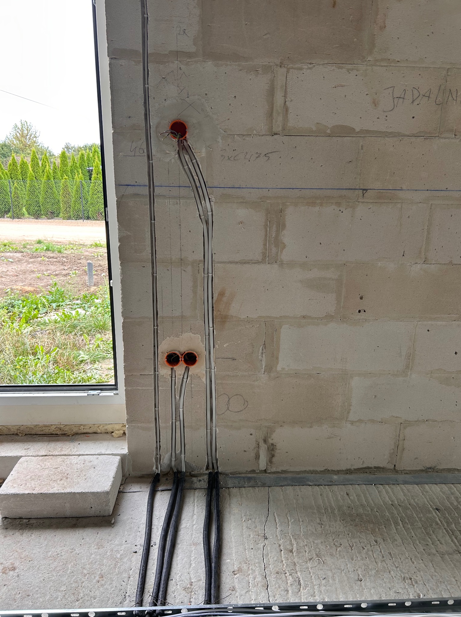 Montaż instalacji elektrycznej w surowym wnętrzu: kable w peszlach prowadzone wzdłuż ściany z betonu komórkowego, widoczne puszki instalacyjne i okno z zielonym tłem.