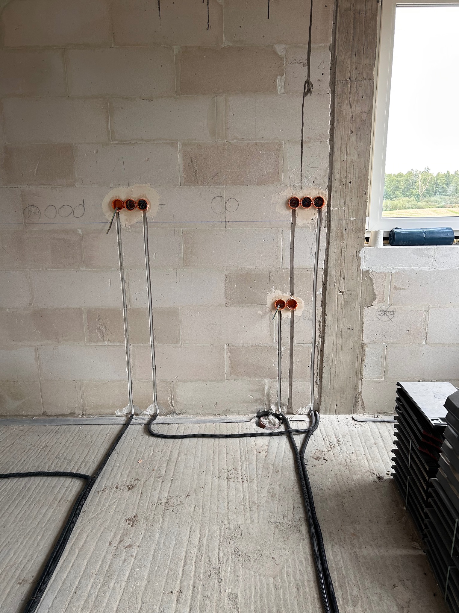 Surowy mur z widocznymi puszkami elektrycznymi i przewodami w szarych rurkach, instalacja w trakcie montażu. Okno w tle.