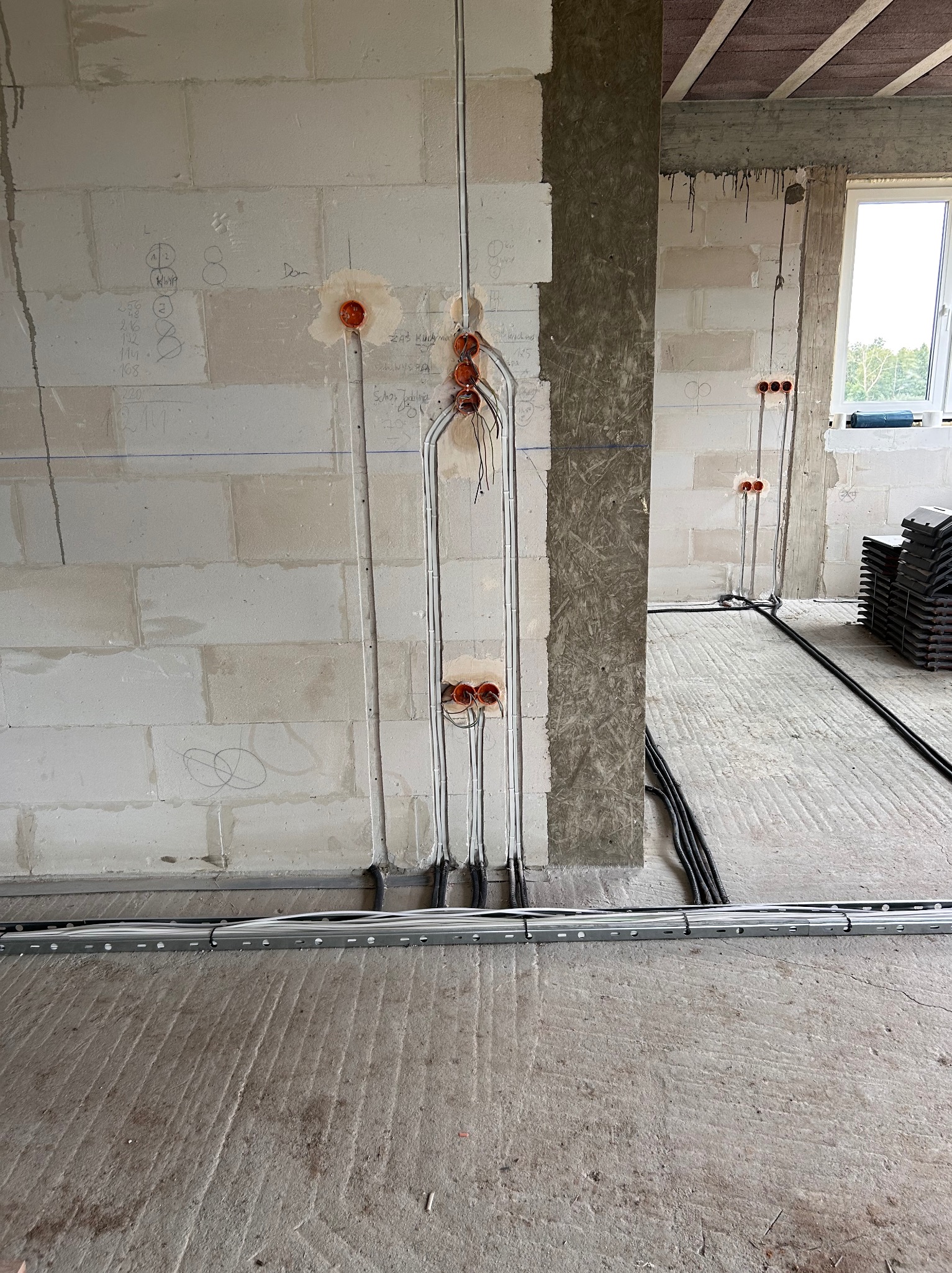 Surowy stan: instalacja przewodów w ścianach z betonu komórkowego i rur osłonowych. Widoczne puszki elektryczne i kable ułożone na podłodze w korytach kablowych.