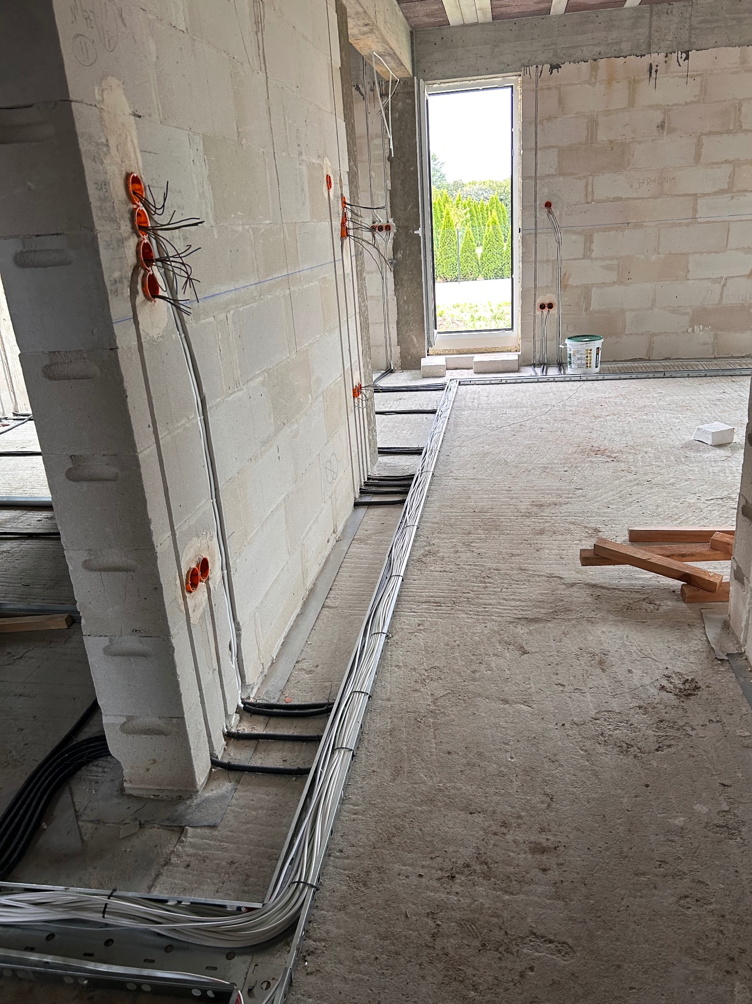 Surowy stan: Instalacja elektryczna w ścianach z betonu komórkowego i koryta kablowe w podłodze, widoczne pomarańczowe puszki i przewody. Okno z widokiem na zieleń w tle.