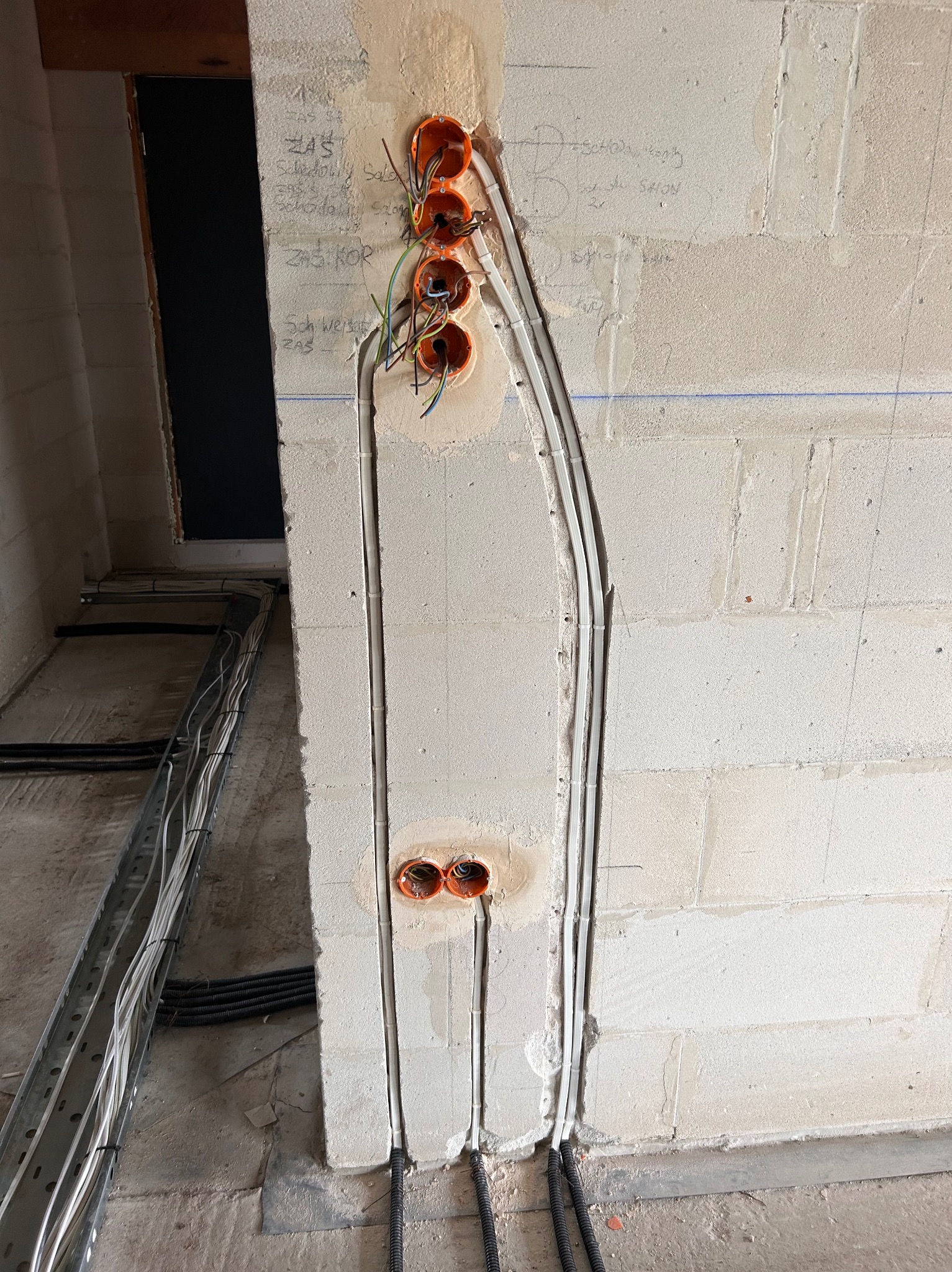 Ściana z pustaków z zamontowanymi puszkami elektrycznymi i przewodami w białych rurkach. Widoczne kable w korytkach na podłodze. Surowy stan wykończenia wnętrza.